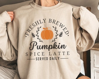 May include: Sand-colored sweatshirt with the text "FRESHLY BREWED. Pumpkin SPICE LATTE SERVED DAILY" in black and orange. The design includes a pumpkin and leaves. The model is wearing a size L.