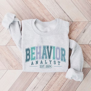 May include: Gray sweatshirt with the text "BEHAVIOR ANALYST EST. 2024" in a colorful, gradient arch design.