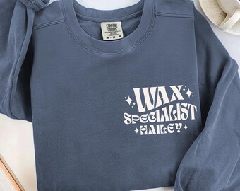 May include: A dusty blue sweatshirt with the text "WAX SPECIALIST HAILEY" in white. The sweatshirt has a crew neck and long sleeves. A coffee cup and a book are in the background.