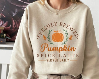 May include: Beige sweatshirt with the text "FRESHLY BREWED. Pumpkin SPICE LATTE SERVED DAILY" in orange and brown. The design includes a pumpkin and leaves. The model is wearing a size M.
