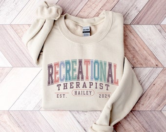 Op de afbeelding: Een beige sweatshirt met de tekst "Recreational Therapist" in regenboogkleuren. De tekst is in een vintage lettertype met de woorden "Est. Hailey 2024" eronder.