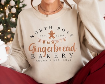 May include: Beige sweatshirt with the text "North Pole Gingerbread Bakery" in brown and black. The design includes a gingerbread man and festive banner. The sweatshirt is part of a holiday-themed collection.