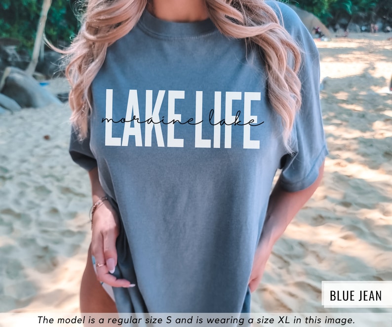 Puede incluir: Una camiseta azul jean con el texto "LAKE LIFE" en letras blancas. El texto "moraine lake" est&aacute; escrito en cursiva debajo del texto principal.