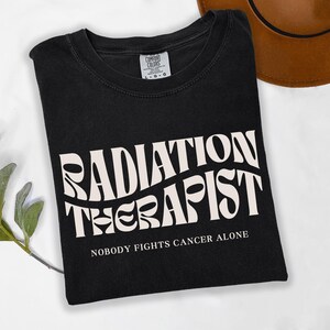 May include: Black t-shirt with white text that reads "Radiation Therapist" in a retro font. Below the text is the phrase "Nobody fights cancer alone."