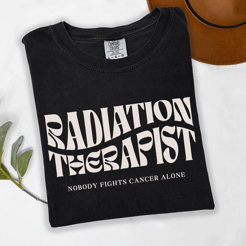 May include: Black t-shirt with white text that reads "Radiation Therapist" in a retro font. Below the text is the phrase "Nobody fights cancer alone."
