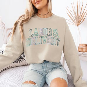 May include: A light beige sweatshirt with the text "LABOR & DELIVERY" in a colorful, varsity-style font.