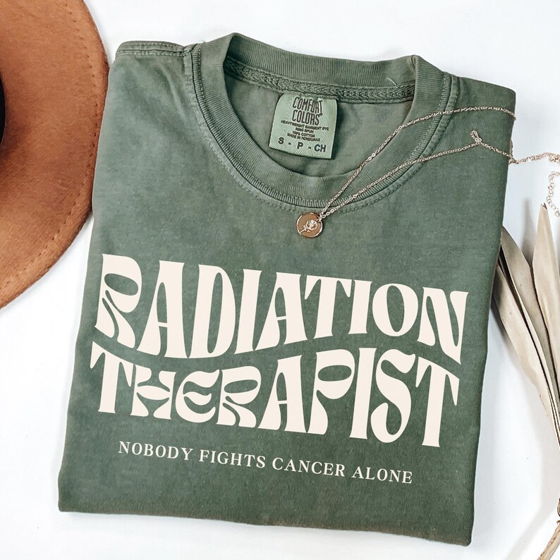 May include: A green t-shirt with the text "RADIATION THERAPIST" in a retro font. The text is in white and the shirt has the words "NOBODY FIGHTS CANCER ALONE" printed below it.