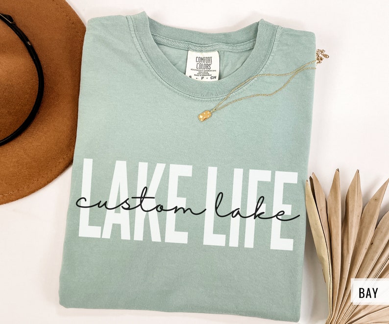Puede incluir: Una camiseta azul claro con el texto "LAKE LIFE" en letras blancas y "custom lake" en letra cursiva negra. La camiseta est&aacute; doblada y colocada sobre una superficie blanca.