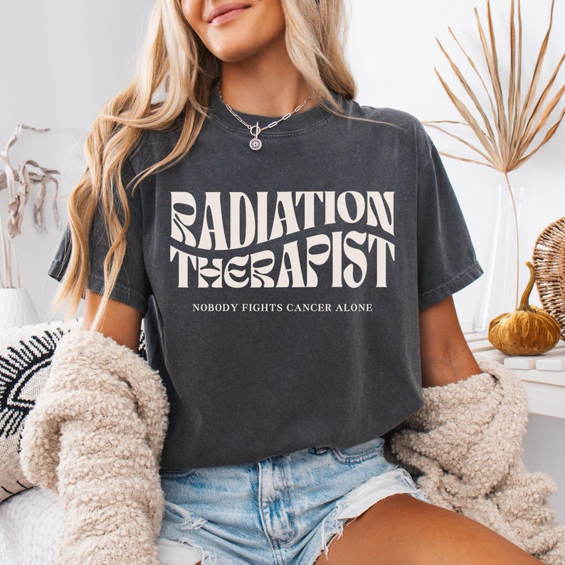 May include: A dark grey t-shirt with a white graphic that reads "Radiation Therapist" in a retro font. The text "Nobody Fights Cancer Alone" is printed below the graphic.