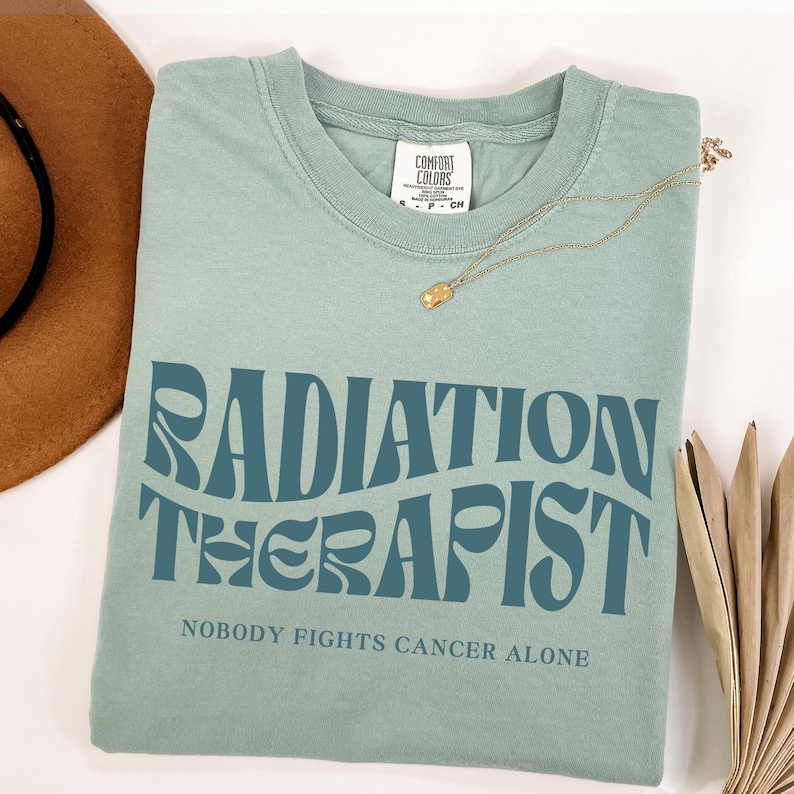 May include: A light blue t-shirt with the text "RADIATION THERAPIST" in a retro font. The text "NOBODY FIGHTS CANCER ALONE" is printed below the main text.