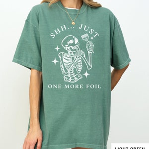 May include: A light green t-shirt featuring a white skeleton graphic holding a hairdressing comb. The text "SHH...JUST ONE MORE FOIL" is printed on the shirt.