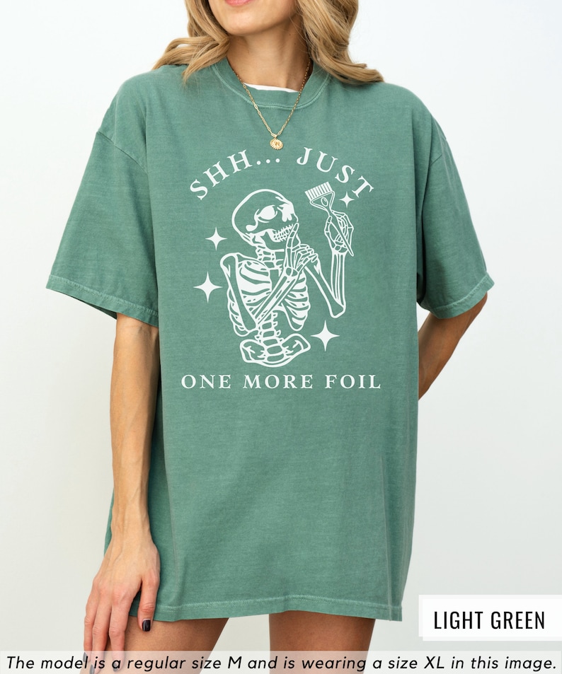 May include: A light green t-shirt featuring a white skeleton graphic holding a hairdressing comb. The text "SHH...JUST ONE MORE FOIL" is printed on the shirt.