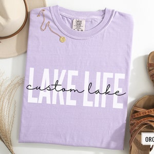 Puede incluir: Una camiseta de color lavanda con el texto "LAKE LIFE custom lake" en letras blancas. El texto est&aacute; en una fuente en negrita y de bloque. La camiseta est&aacute; hecha de un material suave y c&oacute;modo.