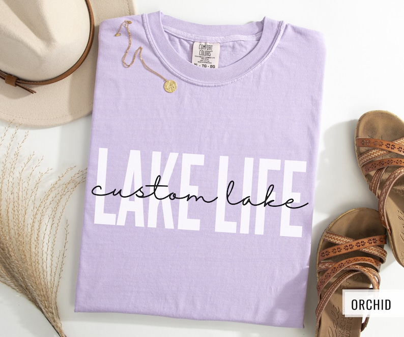 Puede incluir: Una camiseta de color lavanda con el texto "LAKE LIFE custom lake" en letras blancas. El texto est&aacute; en una fuente en negrita y de bloque. La camiseta est&aacute; hecha de un material suave y c&oacute;modo.
