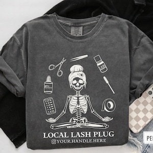 May include: A dark gray t-shirt with a white skeleton wearing a bun and holding lash tools. The text "Local Lash Plug" is printed on the shirt with the social media handle "@your.handle.here".