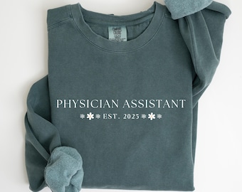 May include: A teal-colored sweatshirt with the words "PHYSICIAN ASSISTANT" in white block letters. Below the text are three snowflakes and the year "EST. 2025". The sleeves are rolled up, and the sweatshirt is made of a soft material.