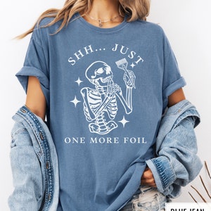 May include: A blue t-shirt featuring a white skeleton holding a hair dye brush with the text "SHH... JUST ONE MORE FOIL".