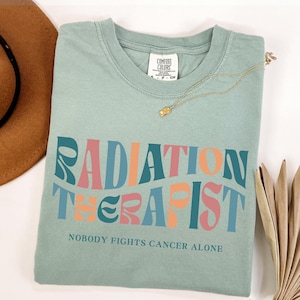 May include: A light blue t-shirt with the text "RADIATION THERAPIST" in a retro font with pink, orange, and blue colors. The text "NOBODY FIGHTS CANCER ALONE" is printed below the main text.