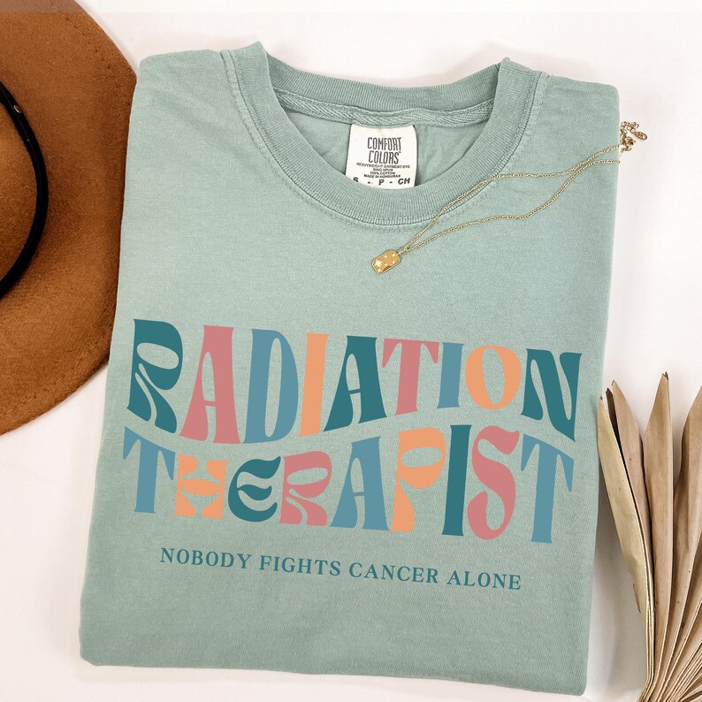 May include: A light blue t-shirt with the text "RADIATION THERAPIST" in a retro font with pink, orange, and blue colors. The text "NOBODY FIGHTS CANCER ALONE" is printed below the main text.