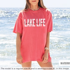 Puede incluir: Una camiseta rosa con el texto "Lake Tahoe Life" en letras blancas. La modelo lleva una talla XL.