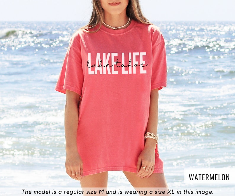 Puede incluir: Una camiseta rosa con el texto "Lake Tahoe Life" en letras blancas. La modelo lleva una talla XL.