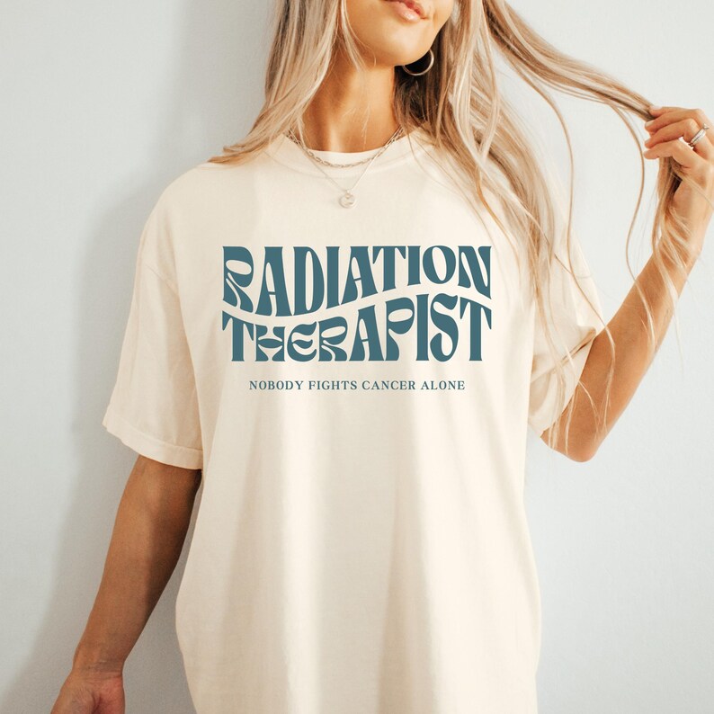 May include: A cream-coloured t-shirt with a blue graphic that says "Radiation Therapist" and "Nobody Fights Cancer Alone".