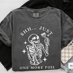 May include: A dark grey t-shirt featuring a skeleton holding a hairdressing comb and the text "Shh... Just One More Foil".