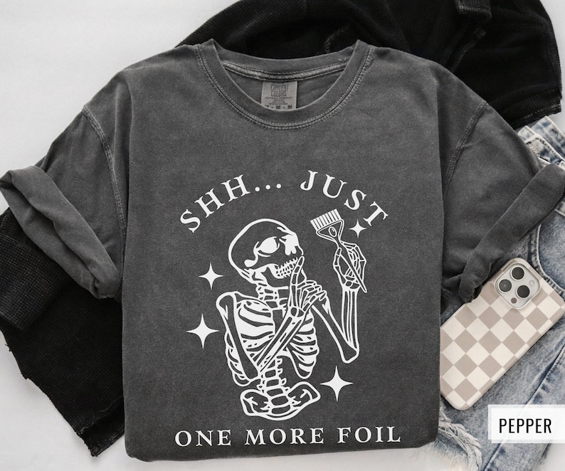 May include: A dark grey t-shirt featuring a skeleton holding a hairdressing comb and the text "Shh... Just One More Foil".