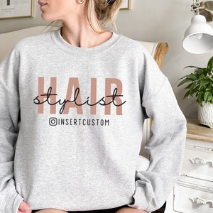 May include: Grey sweatshirt with the text "HAIR stylist" in pink and black lettering. The text "@INSERTCUSTOM" is below the main text in black.