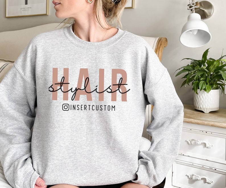 May include: Grey sweatshirt with the text "HAIR stylist" in pink and black lettering. The text "@INSERTCUSTOM" is below the main text in black.