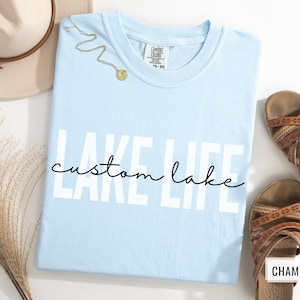 Puede incluir: Camiseta azul claro con el texto "LAKE LIFE" en letras blancas y "custom lake" en escritura cursiva negra debajo. La camiseta est&aacute; colocada sobre una superficie blanca.