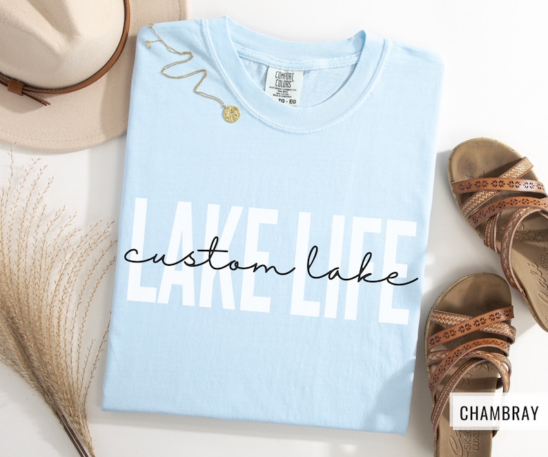 Puede incluir: Camiseta azul claro con el texto "LAKE LIFE" en letras blancas y "custom lake" en escritura cursiva negra debajo. La camiseta est&aacute; colocada sobre una superficie blanca.