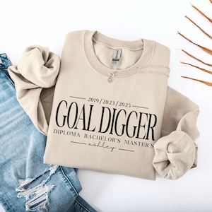 May include: Tan sweatshirt with the text "Goal Digger" and the dates "2019/2023/2025" printed on it. The text "Diploma Bachelor's Master's" is printed below the main text. The name "ashley" is printed below the dates.