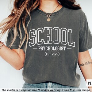 May include: A dark gray t-shirt with white text that reads "SCHOOL PSYCHOLOGIST EST. 2024".
