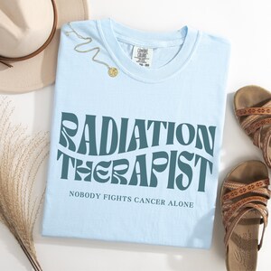 May include: Light blue t-shirt with the text "RADIATION THERAPIST" in a retro font. The text "NOBODY FIGHTS CANCER ALONE" is printed below the main text.