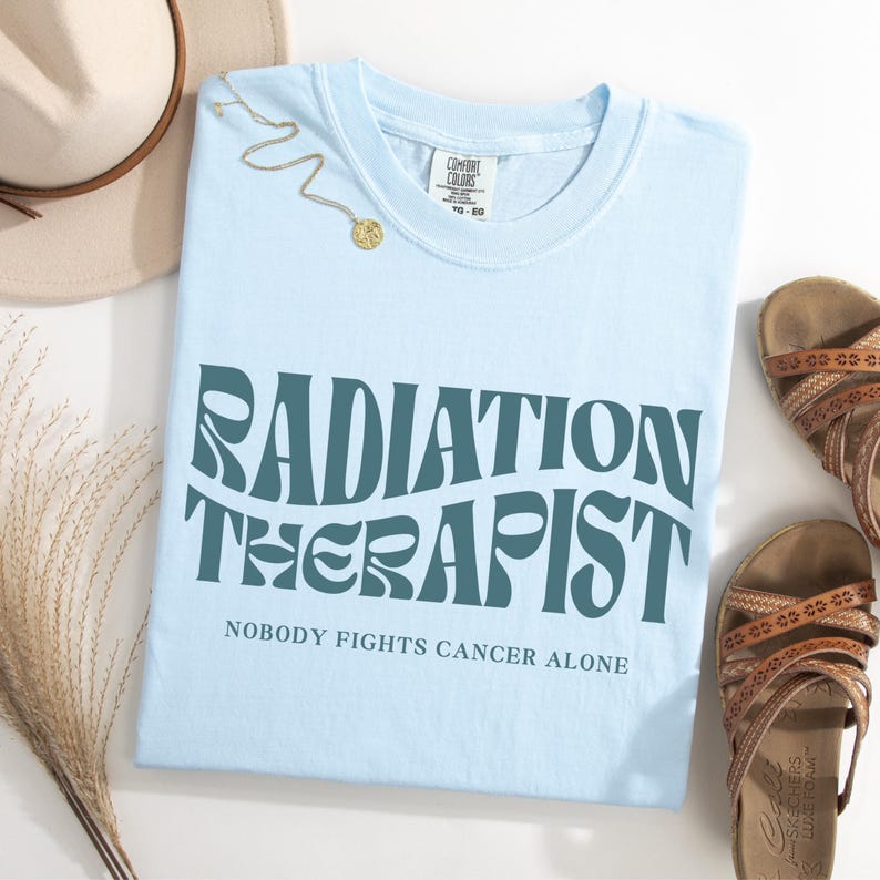 May include: Light blue t-shirt with the text "RADIATION THERAPIST" in a retro font. The text "NOBODY FIGHTS CANCER ALONE" is printed below the main text.