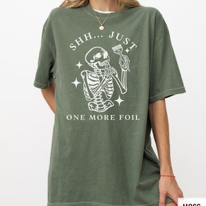 May include: Oversized green t-shirt featuring a white graphic of a skeleton holding a hair dye brush and the text "SHH... JUST ONE MORE FOIL".