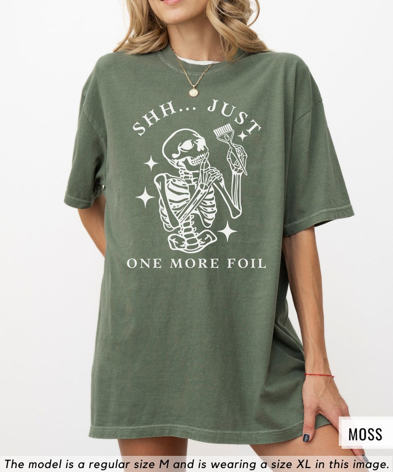 May include: Oversized green t-shirt featuring a white graphic of a skeleton holding a hair dye brush and the text "SHH... JUST ONE MORE FOIL".