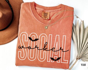 May include: Coral-colored t-shirt with the words "SOCIAL worker" in white, with bat graphics. A gold necklace and a brown hat are visible. The shirt is made of Comfort Colors material.