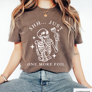 May include: A brown t-shirt featuring a white skeleton graphic holding a comb and scissors. The text "SHH... JUST ONE MORE FOIL" is printed on the shirt.