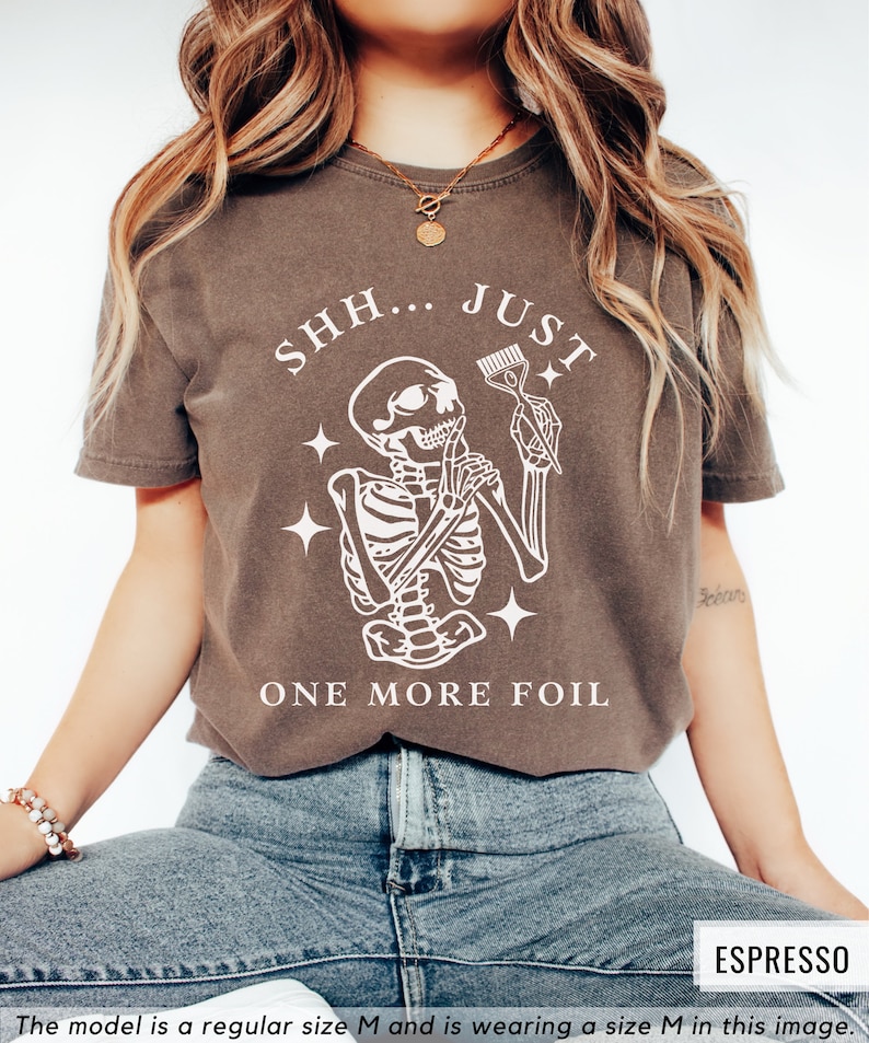 May include: A brown t-shirt featuring a white skeleton graphic holding a comb and scissors. The text "SHH... JUST ONE MORE FOIL" is printed on the shirt.