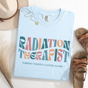 May include: Light blue t-shirt with the text "RADIATION THERAPIST" in a retro font with colorful accents. The text "NOBODY FIGHTS CANCER ALONE" is printed below the main text.