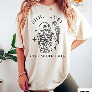 May include: A beige t-shirt featuring a skeleton holding a hair dye brush with the text "SHH... JUST ONE MORE FOIL".