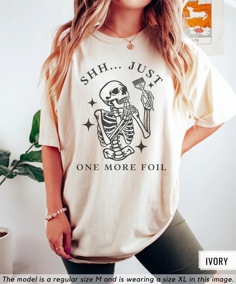 May include: A beige t-shirt featuring a skeleton holding a hair dye brush with the text "SHH... JUST ONE MORE FOIL".