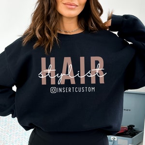 May include: Black sweatshirt with the text "HAIR stylist" in pink and white lettering. The text "@INSERTCUSTOM" is below the main text.