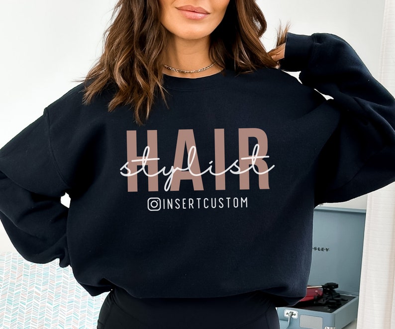 May include: Black sweatshirt with the text "HAIR stylist" in pink and white lettering. The text "@INSERTCUSTOM" is below the main text.