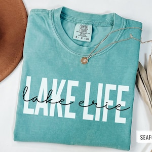 Puede incluir: Una camiseta azul turquesa con el texto "lake erie" en letras blancas y "lake" en letra cursiva negra. La camiseta est&aacute; doblada y tiene una etiqueta que dice "Comfort Colors" y "S - P - CH". El fondo es blanco.