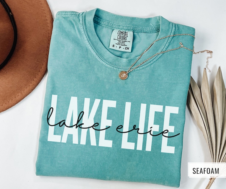 Puede incluir: Una camiseta azul turquesa con el texto "lake erie" en letras blancas y "lake" en letra cursiva negra. La camiseta est&aacute; doblada y tiene una etiqueta que dice "Comfort Colors" y "S - P - CH". El fondo es blanco.