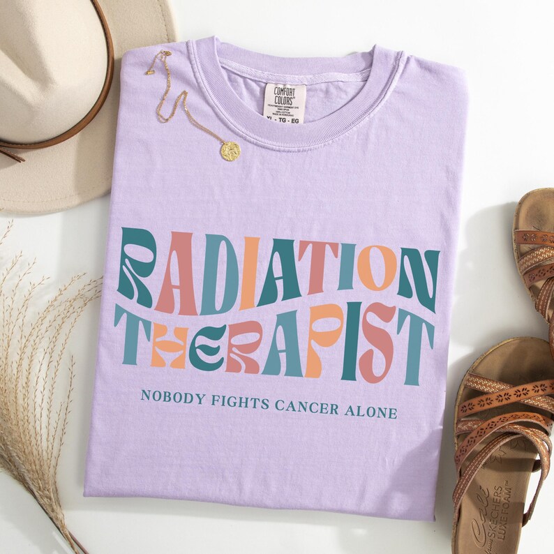 May include: Purple t-shirt with the text "Radiation Therapist" in a retro font with pink, orange, and teal colors. The text "Nobody Fights Cancer Alone" is printed below the main text.