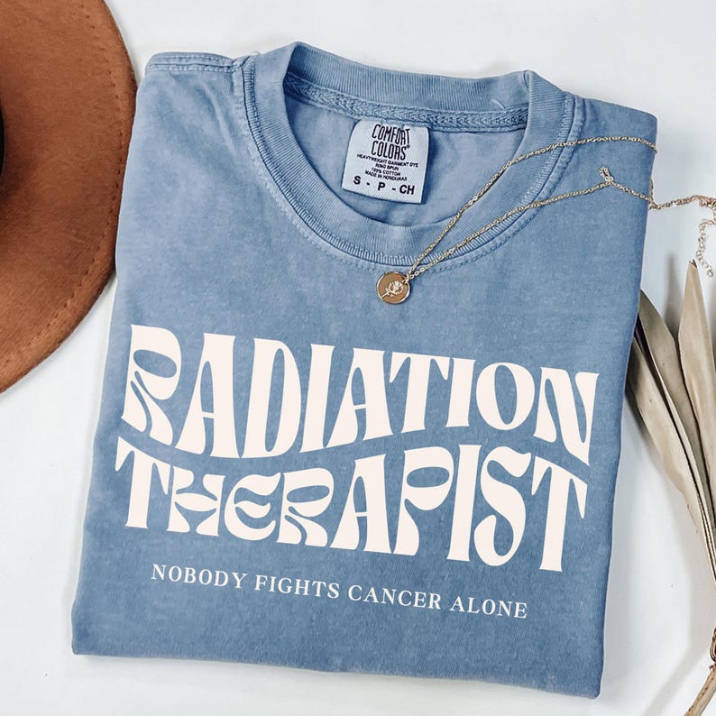May include: A light blue t-shirt with a white graphic that says "Radiation Therapist" in a retro font. The text below the graphic says "Nobody Fights Cancer Alone".
